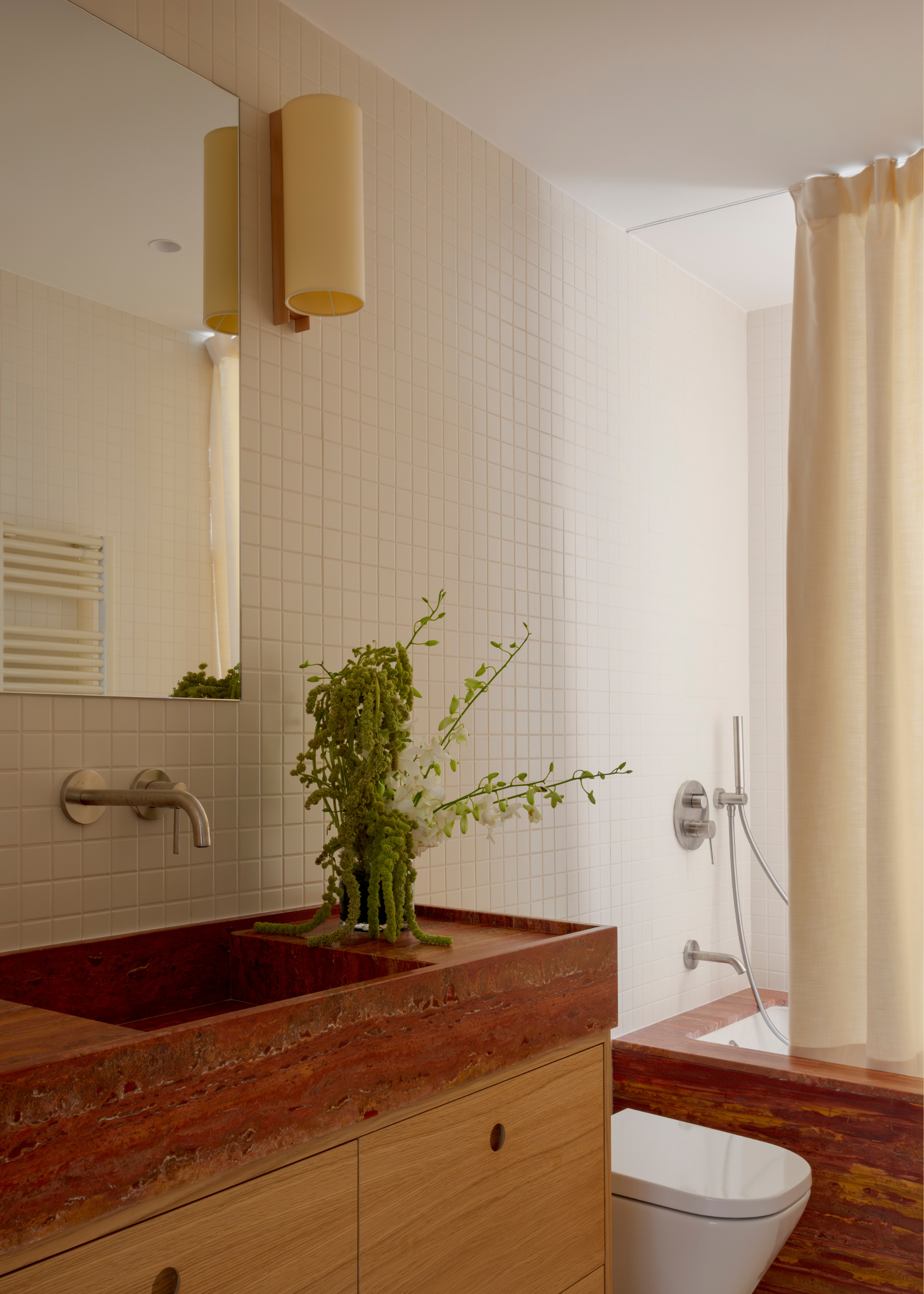 A white tiled bathroom with a stone sink, a green stem arrangement, a wall lamp, a mirror, and a tub area with a shower