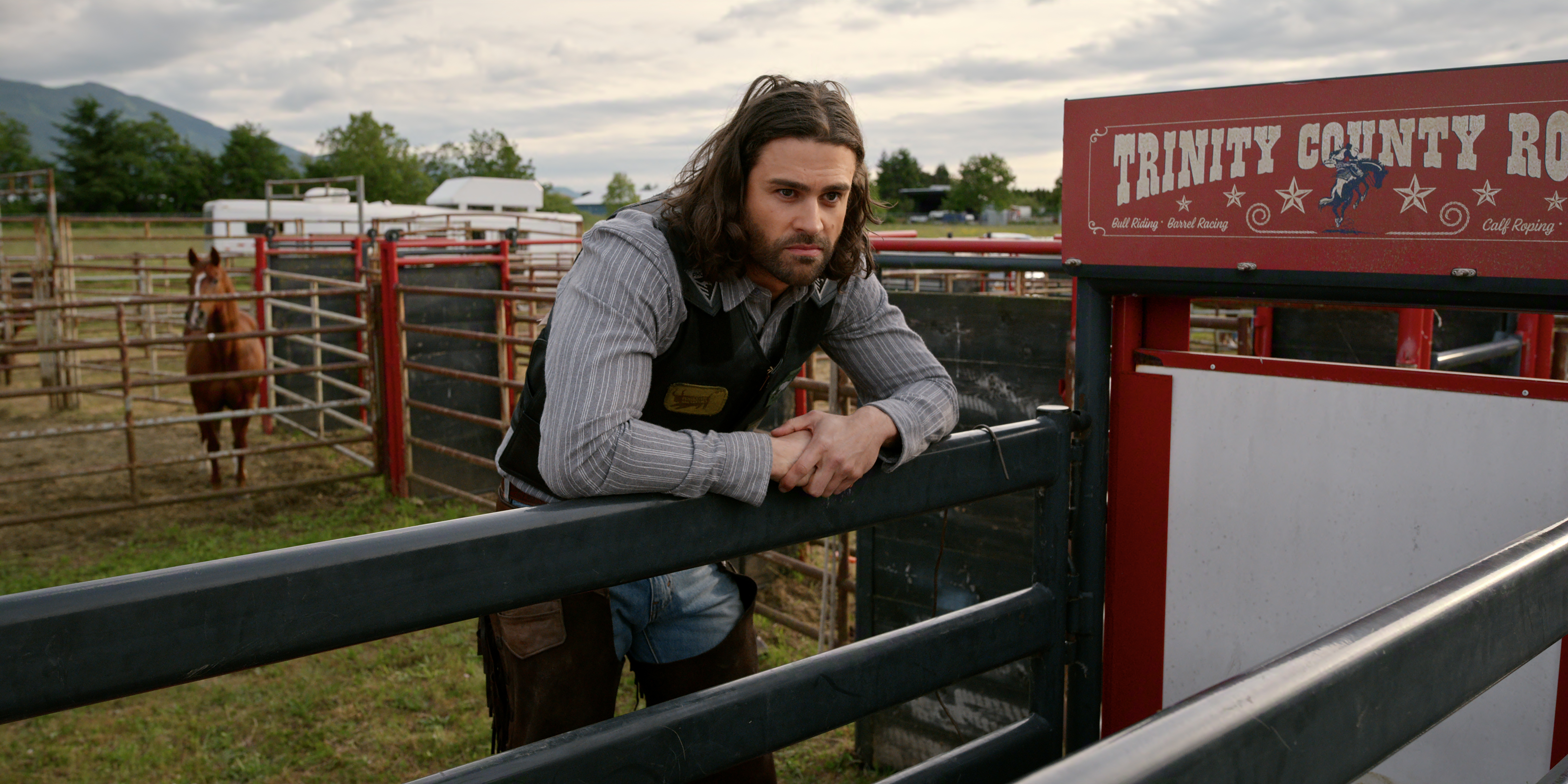 Cody Kearsley as Clay in Episode #708 of Virgin River Season 7 wearing western wear leaning over a fence with horses behind him