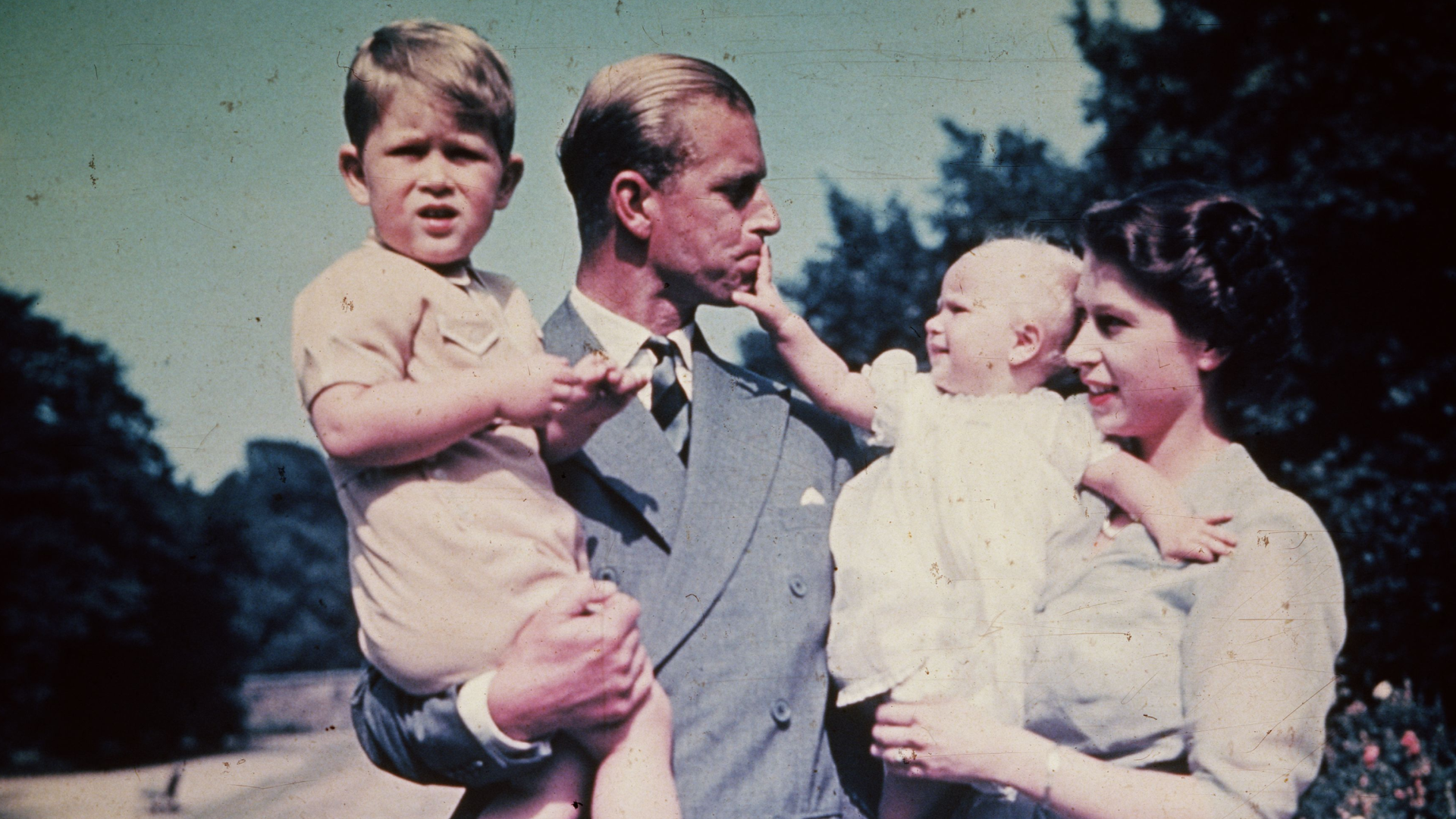 Prince Philip holding Prince Charles while Princess Anne, held by Queen Elizabeth, pulls on his lip 