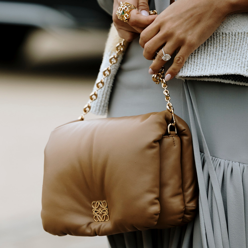 A close up photograph of a woman's hands, the woman wears an elegant diamond ring, carries a Loewe cloud bag and has an oval shaped manicure with light pink nail polish.