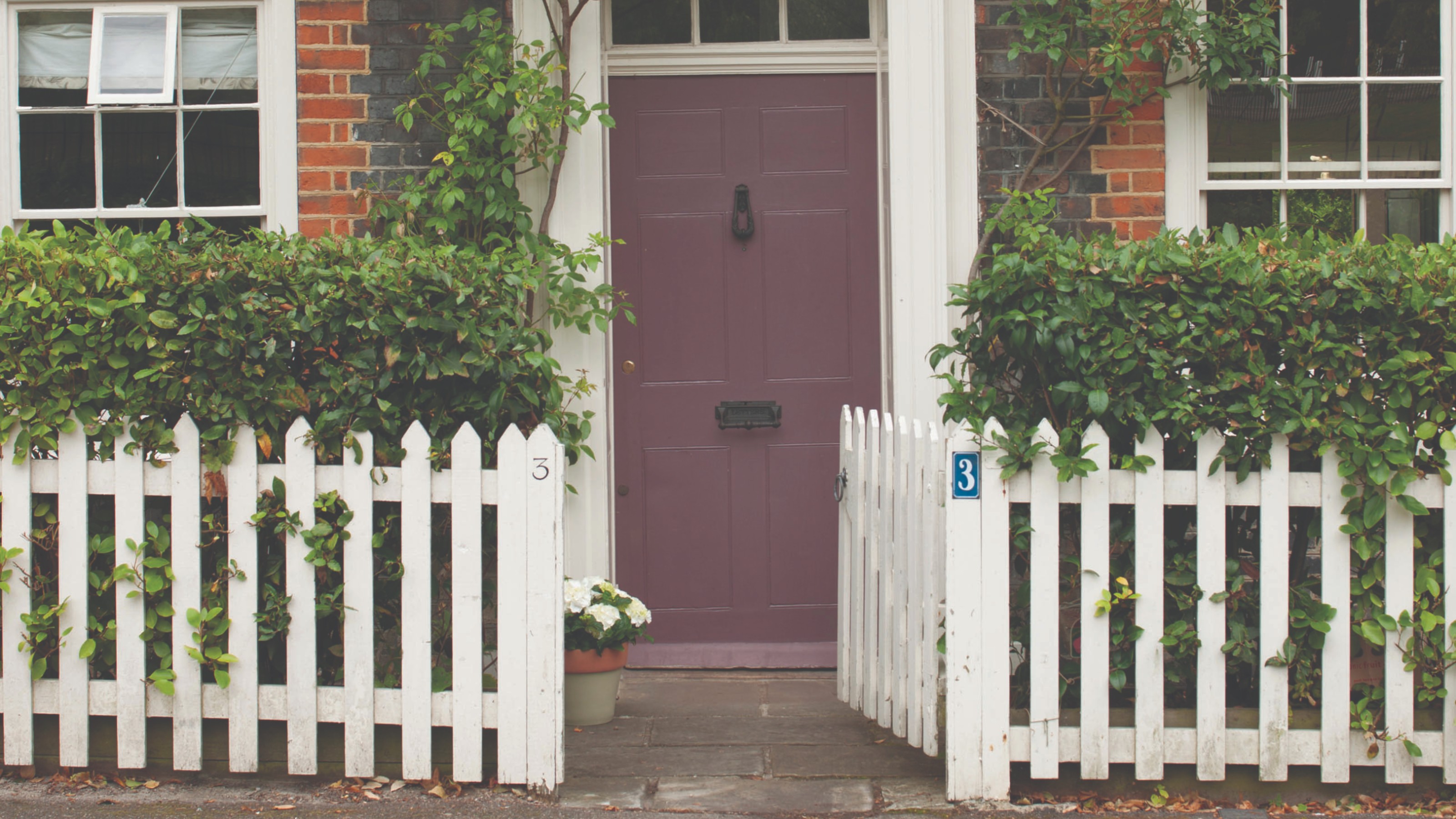 A front door of a red brick house painted in Farrow &amp; Ball's Brassica shade with a white face in front of it