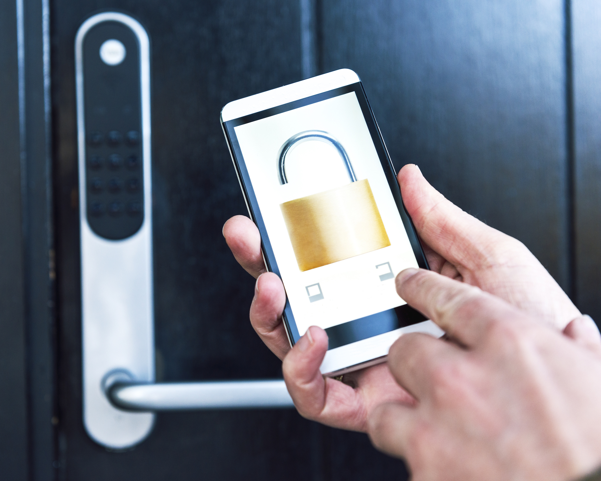 Hands holding smartphone with padlock image on screen in front of black front door with smart lock