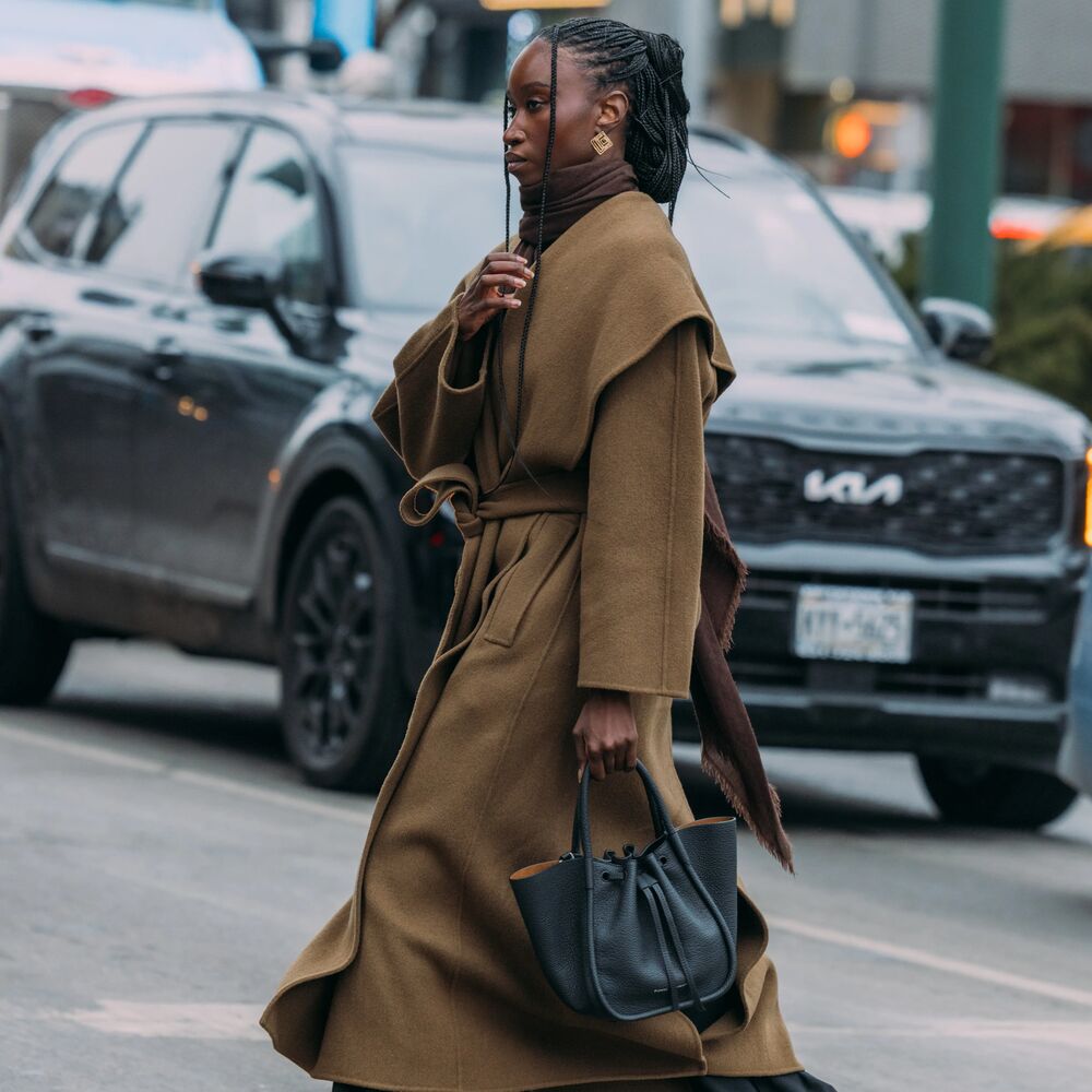 Woman wears long brown coat, black pants, loafers, black handbag.