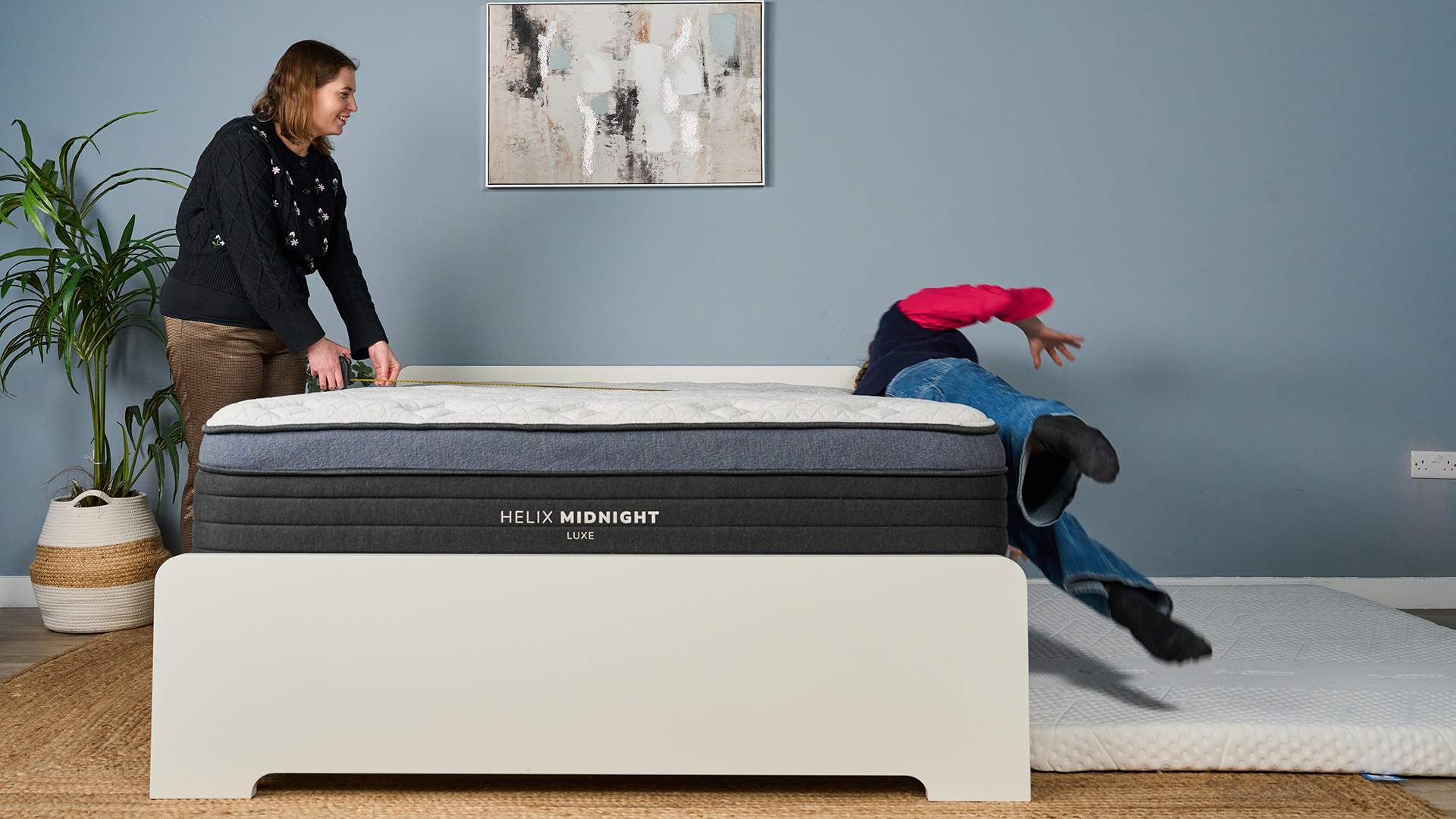 Mattress tester Nicola Appleton rolls of the side of the Helix Midnight Luxe mattress in our Sleep Studio, as she tests the edge support. Tester Ruth Jones stands on the other side of the other side of the mattress, measuring the space on the surface