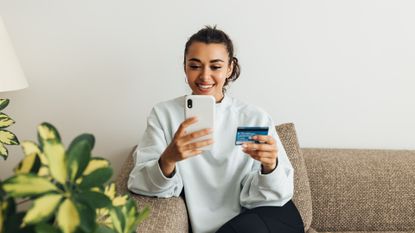 Smiling Woman Holding Credit Card While Using Smart Phone At Home 