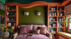A living room with green wallpaper-ed and painted walls holding orange book shelves with colorful books, ceramics and vases. Pastel glass sconces sit on the wall above a dusty pink couch with trimmed pillows and vases of flowers are on either side of the room