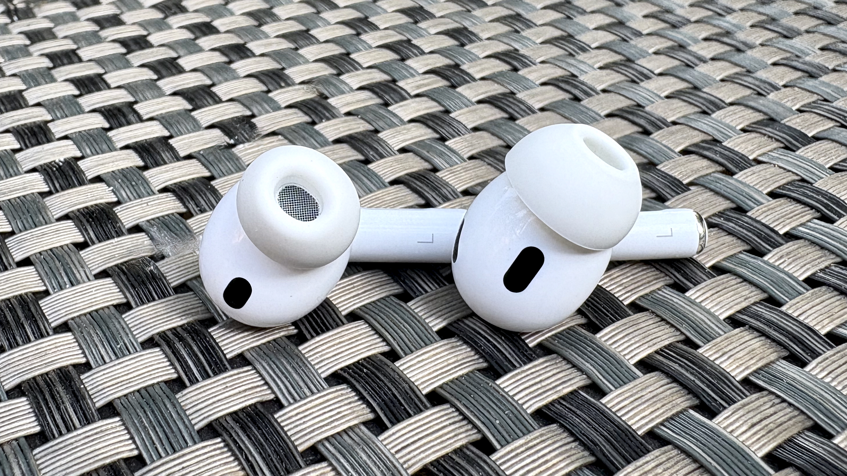 Apple AirPods Pro 3 either worn by a man standing in front of a red-brick wall, or on a table with gray twine cover