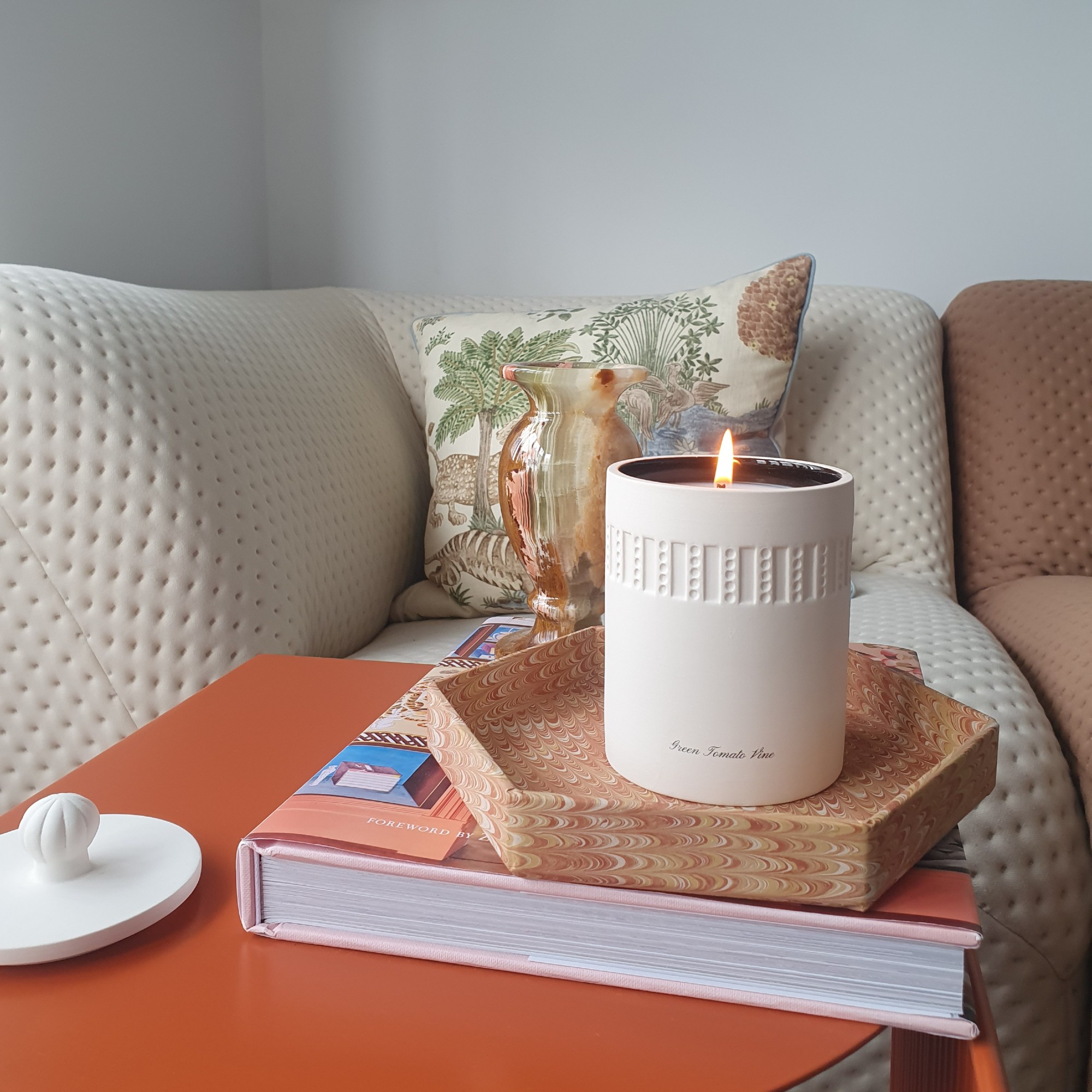 A lit Jo Malone Green Tomato Vine candle on an orange coffee table with a coffee table book