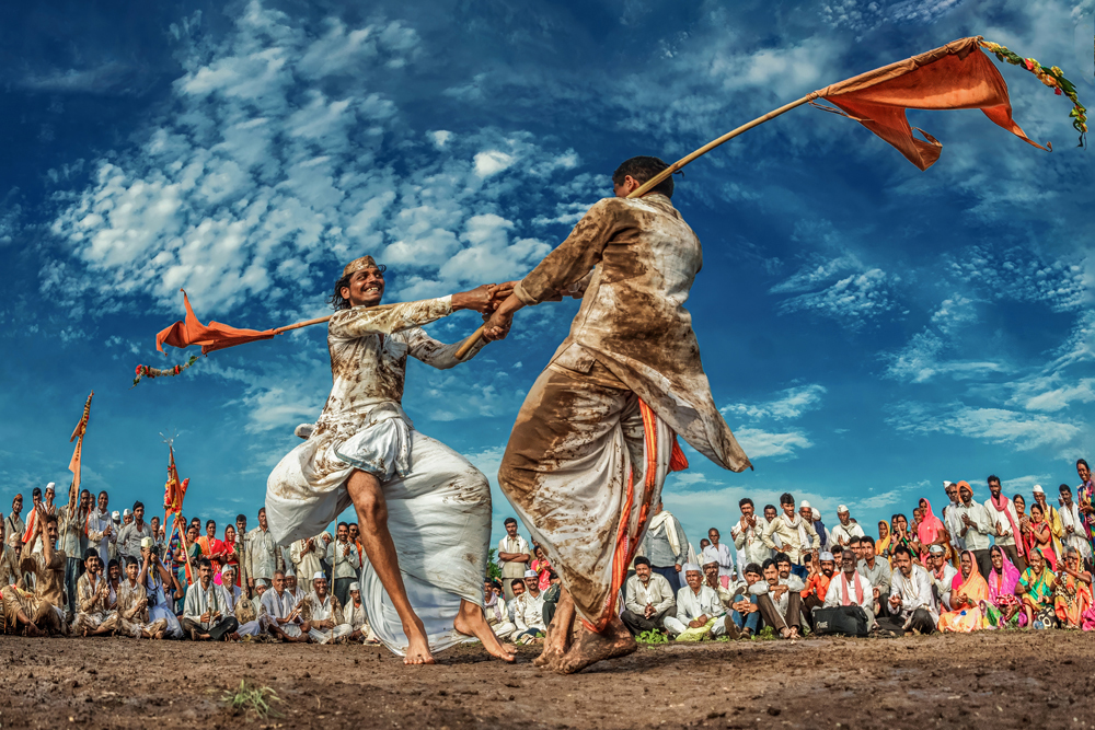 Two men in traditional attire engage in a vigorous dance with flags, surrounded by an audience in colorful clothing under a vibrant sky