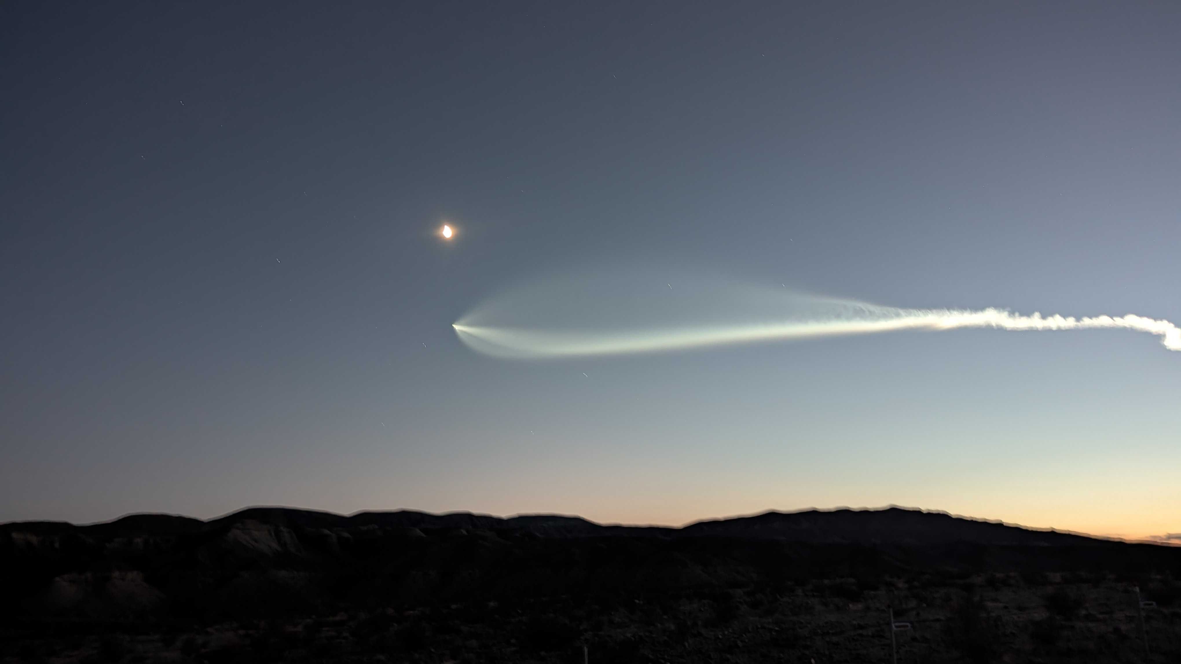 an expanding white plume extends from a small streak of white, growing thicker in a stream across the darkening blue sky below a small shining moon.