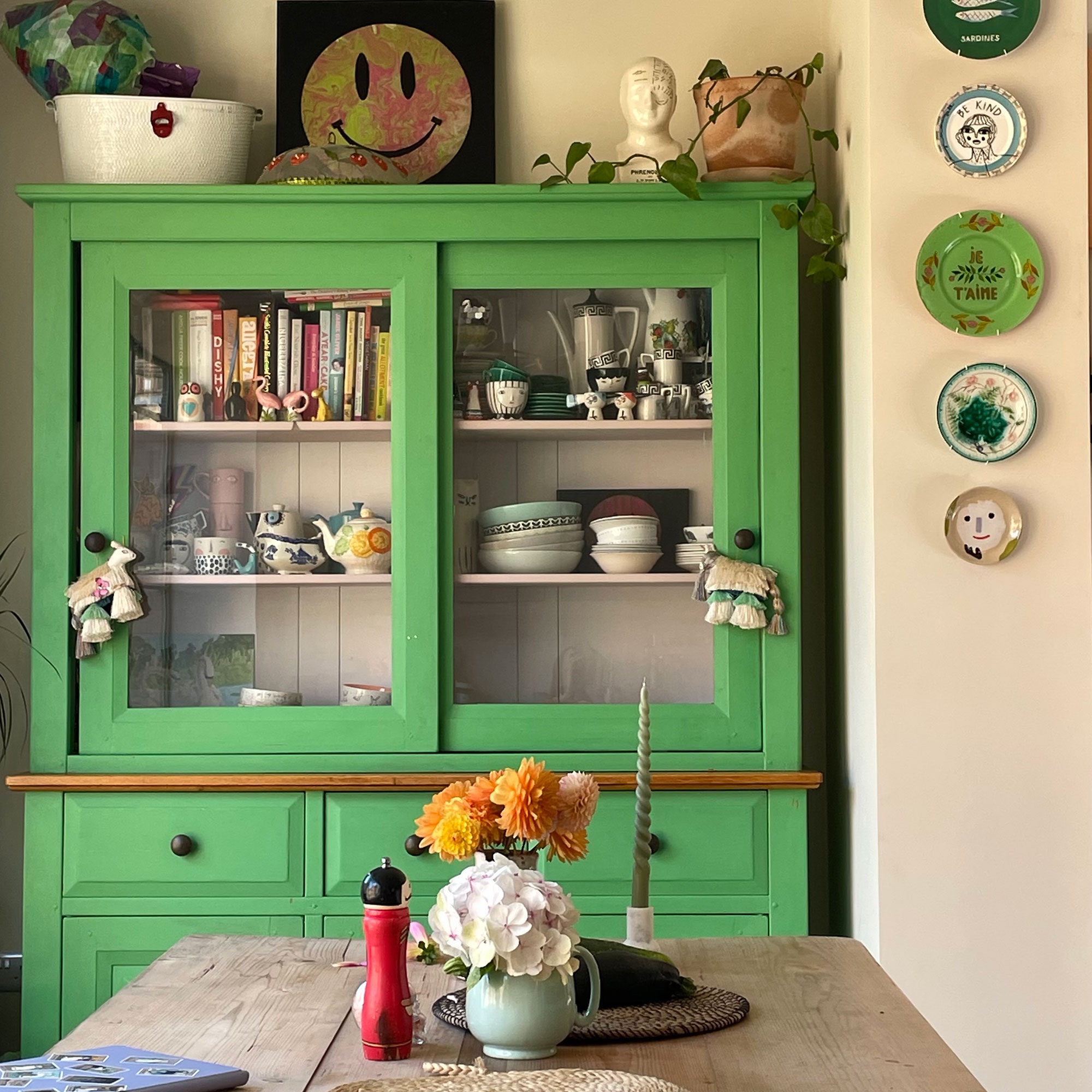 Glass-fronted, green painted wooden dressed holding cookbooks and crockery beside plate display on wall