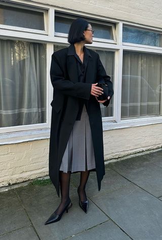 winter tight outfit shown in a photo of a woman wearing a black coat over a black shirt with a gray pleated skirt, black tights, and black pumps