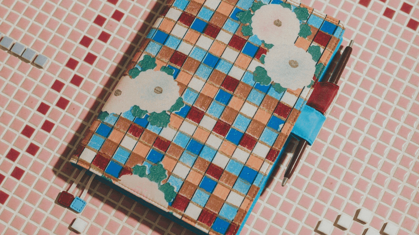 Rotating gif showcasing a checkered Hobonichi Cover Cousin (A5) Unsodo Shin-Bijutsukai (Flower) A5 Cousin Size 2, decorated with blue, white, red, and brown squares and flowers, sitting against a checkered pink, white, and red background.