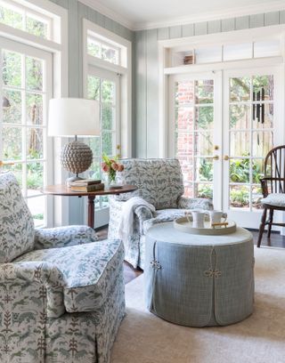 living room blue patterned armchairs with pale blue paneled walls and french windows to garden and round pouffe with mugs on tray and dark wood round pedestal lamp table and dark wood windsor chair