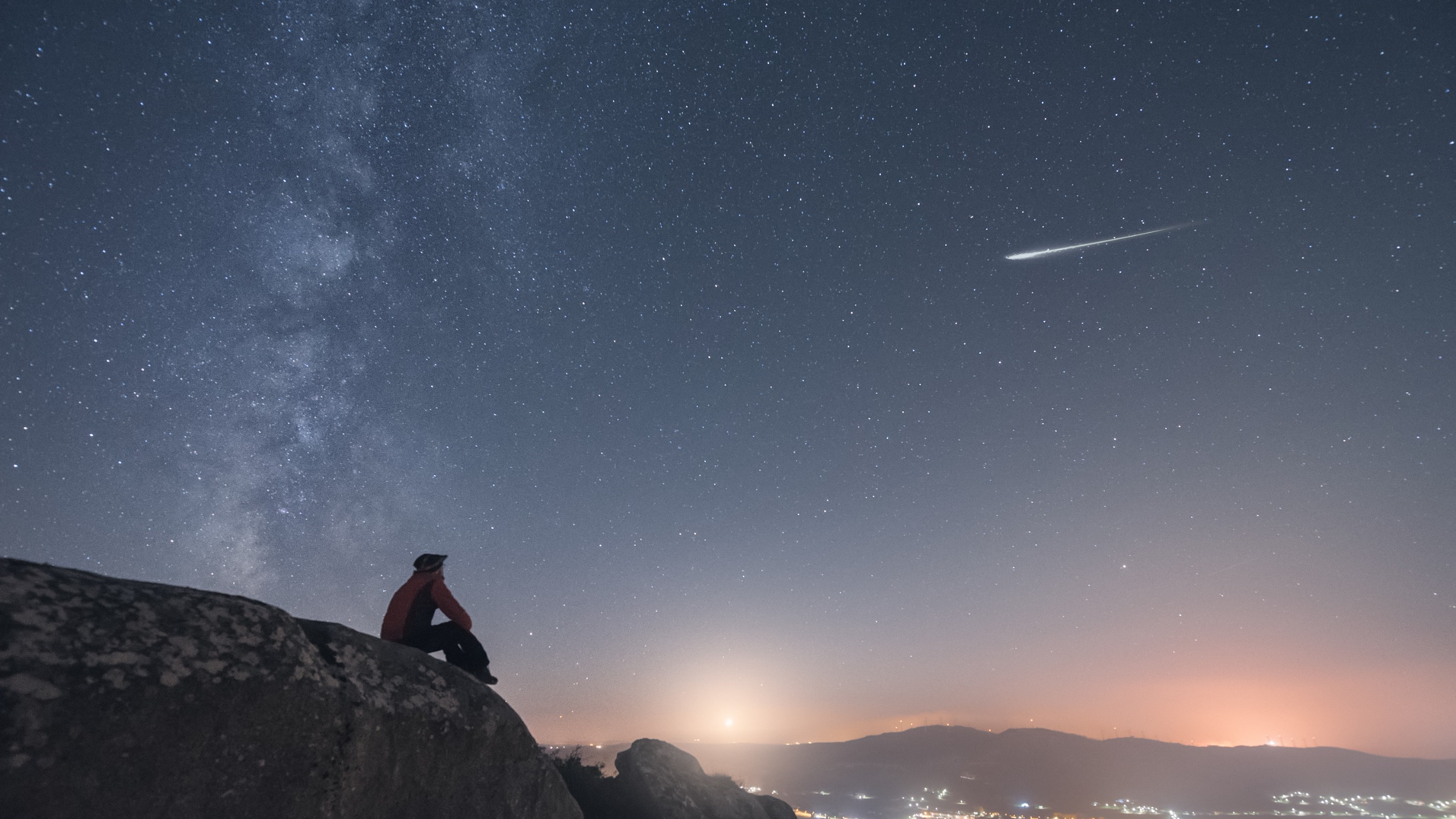 a person sits looking out at a starry sky with the milky way on the left and a meteor streaking through the sky on the right.