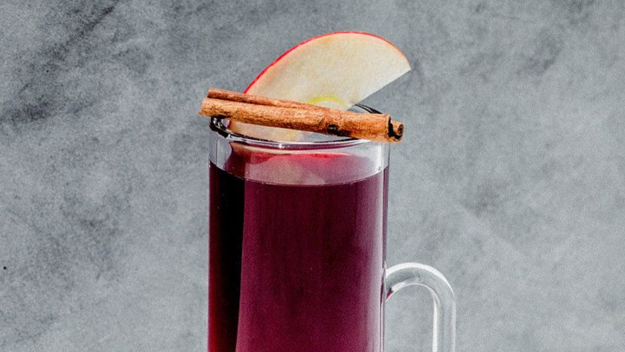 a garnet-colored liquid fills a tall tumbler. it is garnished with a cinnamon stick and a thin apple slice