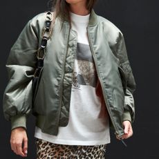 Model wearing a khaki bomber jacket, white graphic t-shirt and leopard print trousers with a handbag all sold at Coggles