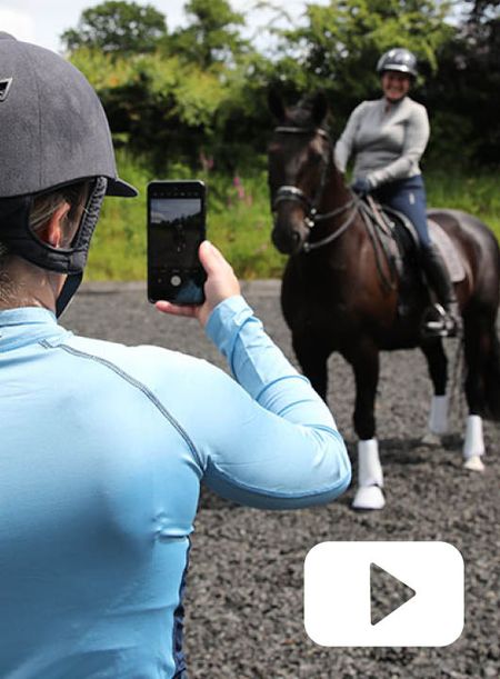A person wearing a riding hat hold up a mobile phone to video a horse and rider who are dressed for dressage schooling at home. 
