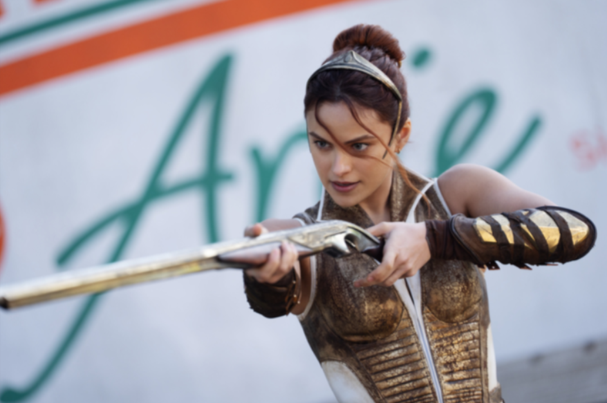Uma mulher usando uma coroa e uma armadura de bronze aponta para um rifle.
