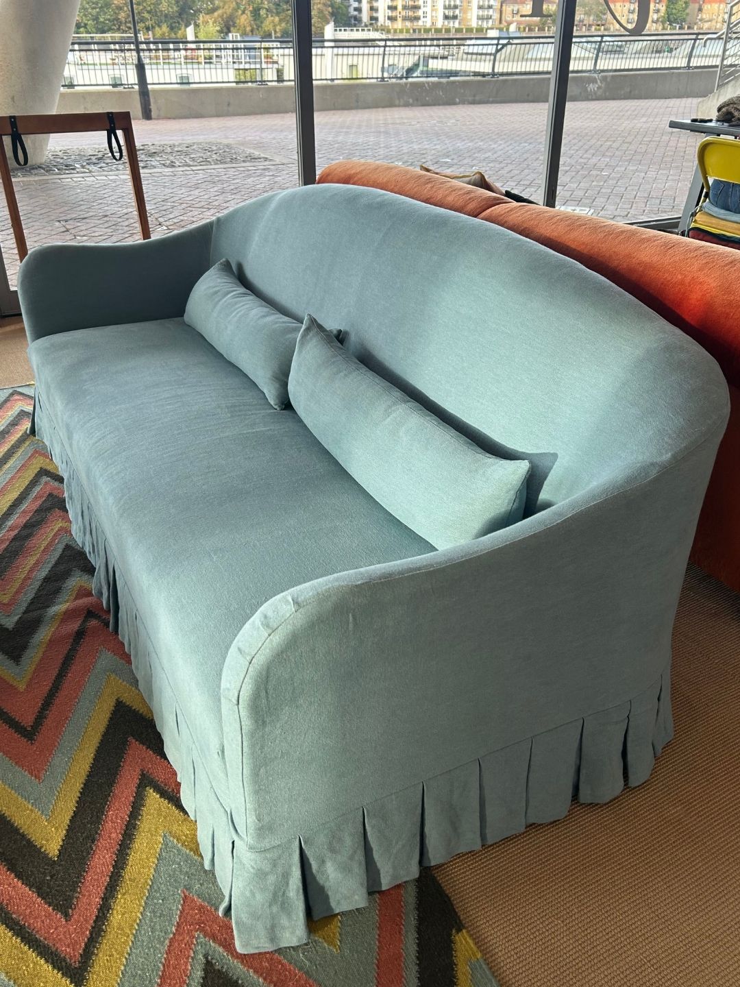 Schplendid's Edie sofa upholstered in pale blue Skye linen, featuring a curved camelback, two bolster cushions, rounded arms and a tailored pleated skirt.