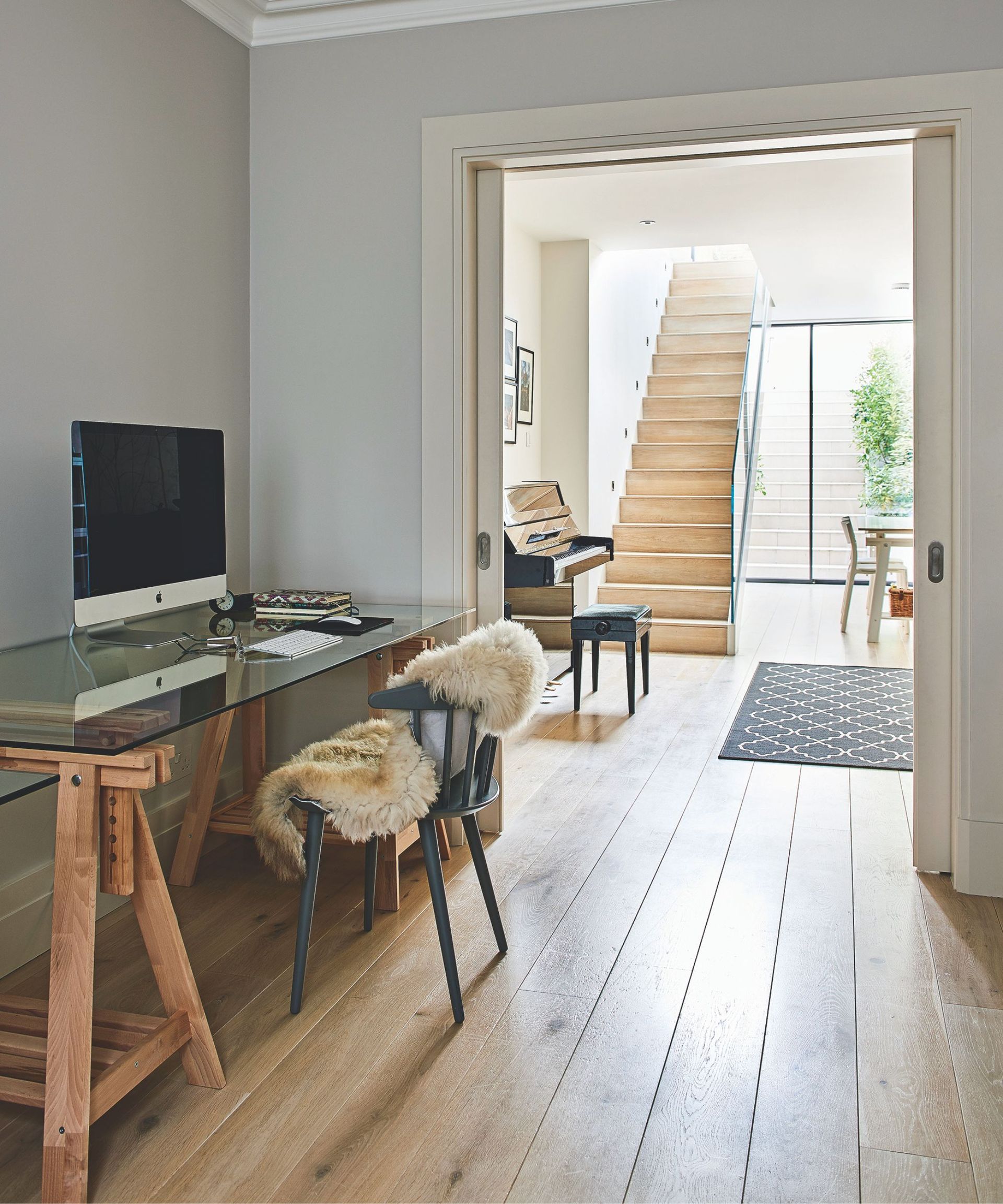 A bright and airy basement with wooden flooring, a wooden and glass desk with a wooden chair with a fluffy throw, and a piano in the background adjoining room with a black patterned rug and wooden staircase upstairs. In the background are glass doors out to a garden staircase, with a leafy green plant to the right.