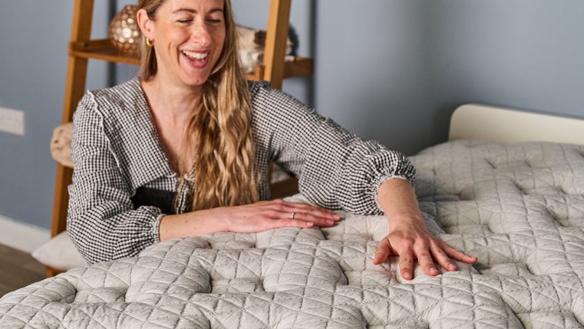 The image shows an woman smiling as she places her hands on the surface of a white mattress while she crouches next to it