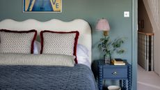 Cozy bedroom with a scalloped white headboard, blue velvet quilt, patterned throw pillows, and a blue bobbin nightstand with a pleated lamp against a sage green wall.