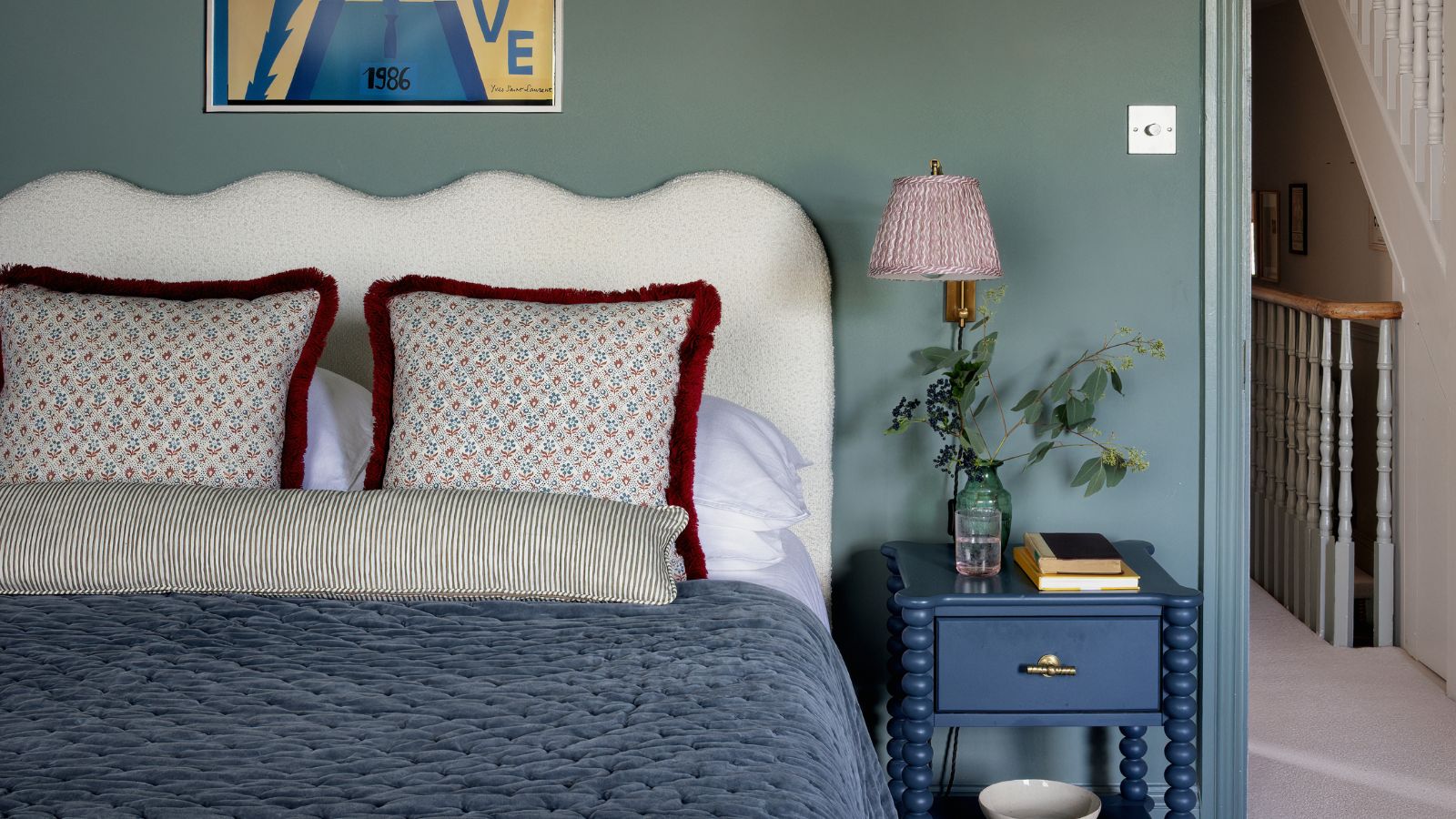Cozy bedroom with a scalloped white headboard, blue velvet quilt, patterned throw pillows, and a blue bobbin nightstand with a pleated lamp against a sage green wall.