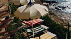 Image from above of several outdoor loungers on a stone area by the ocean. There are yellow, green, and red striped towels on the chairs and white parasols. 