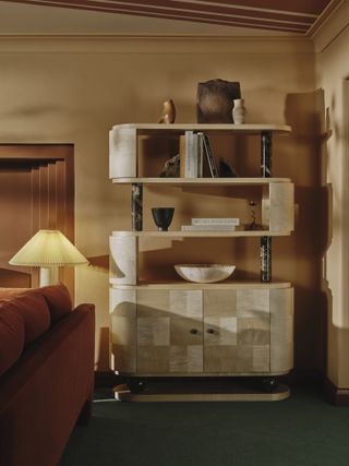 Image of the back corner of a living room. There is a bookshelf cabinet that is made of white wood material in a checkerboard cabinet and has green marble pillars. There are various bowls, vases, and books stacked on the four shelves. The carpet is a green jewel tone, the walls are a beige color, and the sofa is a burnt orange. There is a fluted lampshade by the sofa.