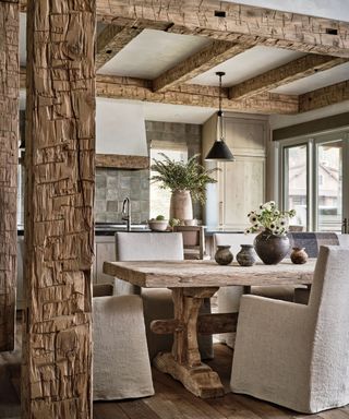Traditional kitchen-dining room with rustic ceiling beams and pillars, dark wood flooring, neutral colored space, large, rustic wood dining table, dining chairs with fabric seat coverings, table decorated with vases, vase of flowers in background on kitchen island