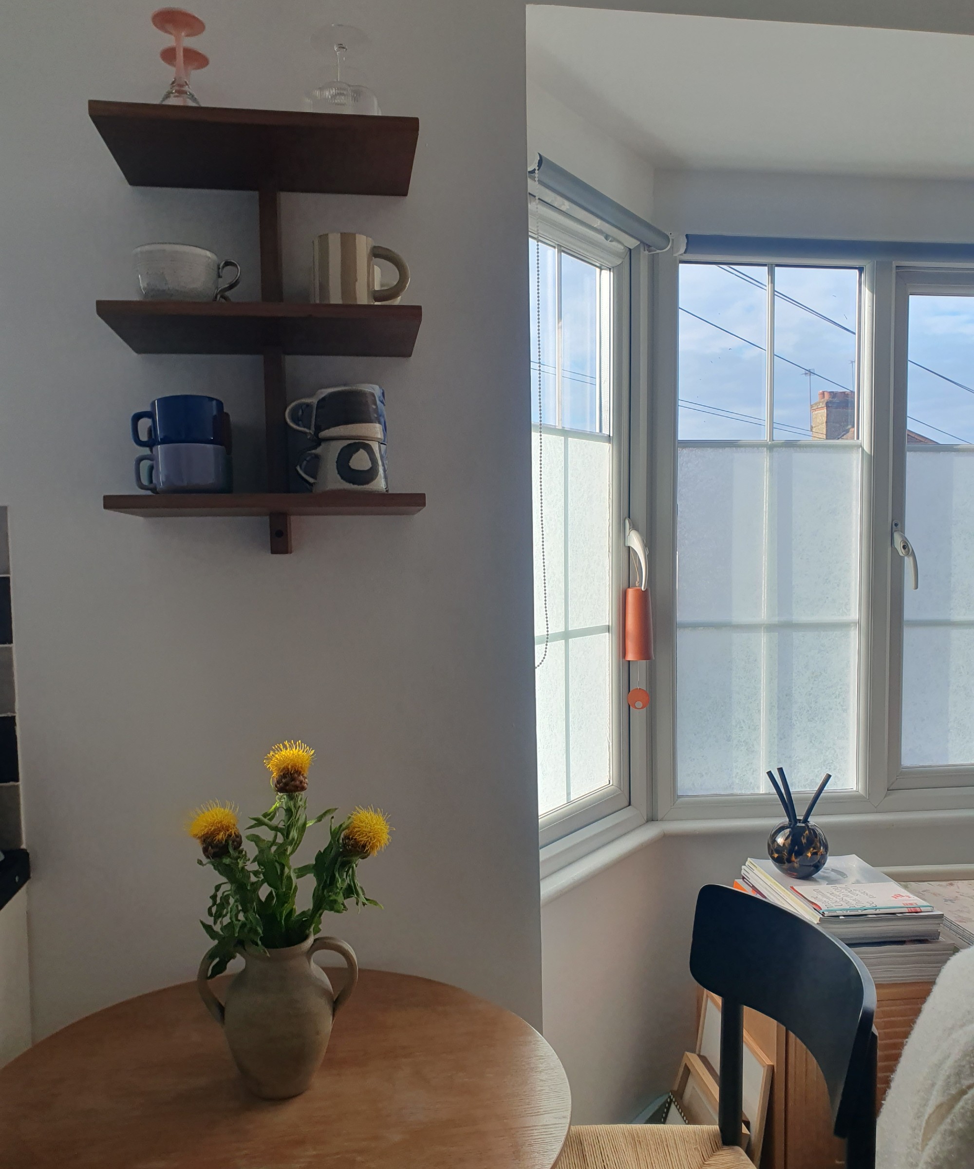 A small round dining table with a vase of flowers and a wall shelf above it displaying mugs