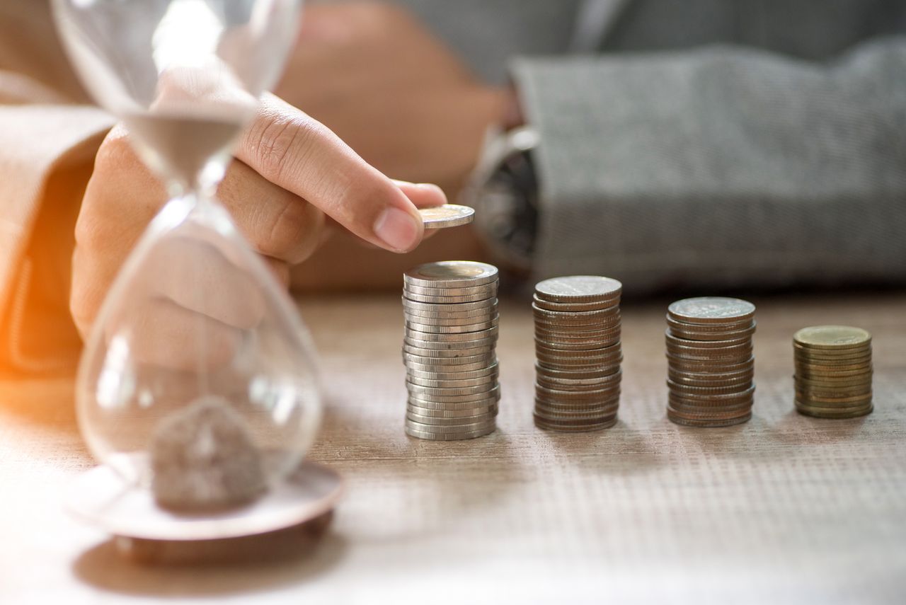 A man sits behind a table with an hourglass and coins on it, symbolising the long wait for Woodford compensation. 