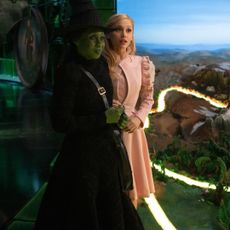 Cynthia Erivo as Elphaba and Ariana Grande as Glinda, standing on the Wicked Part One set next to a miniature of the Yellow Brick Road