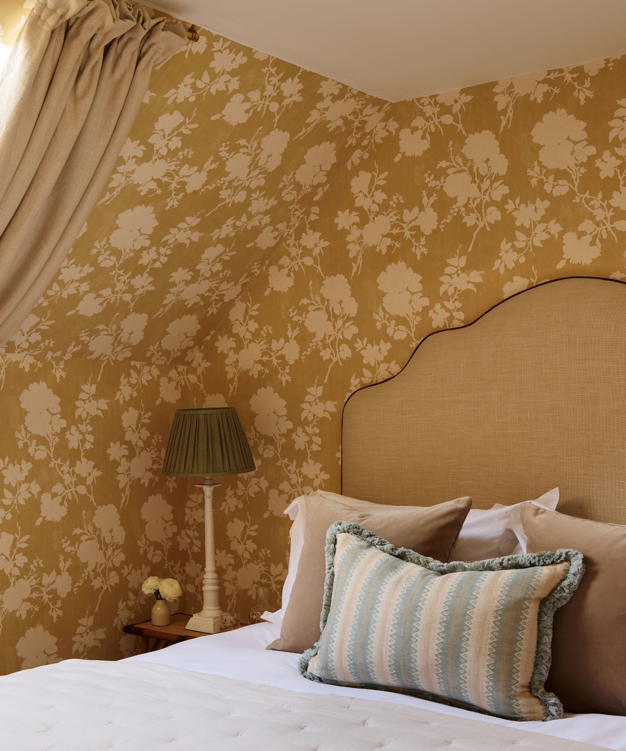 Cozy attic bedroom featuring sloped ceilings covered in mustard-yellow floral wallpaper, a tall beige headboard, and soft lighting