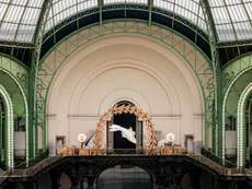 A gigantic fish-shaped sculptures floats in the air under a wooden arch in a green-hued, stunning glasshouse-like building.