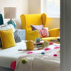 Yellow armchair behind bed with stack of books and yellow teapot resting on top