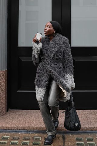 A woman standing on the sidewalk wearing a black headband, silver earrings, a gray fur coat, gray leather pants, a black Prada tote bag, and black ankle boots.