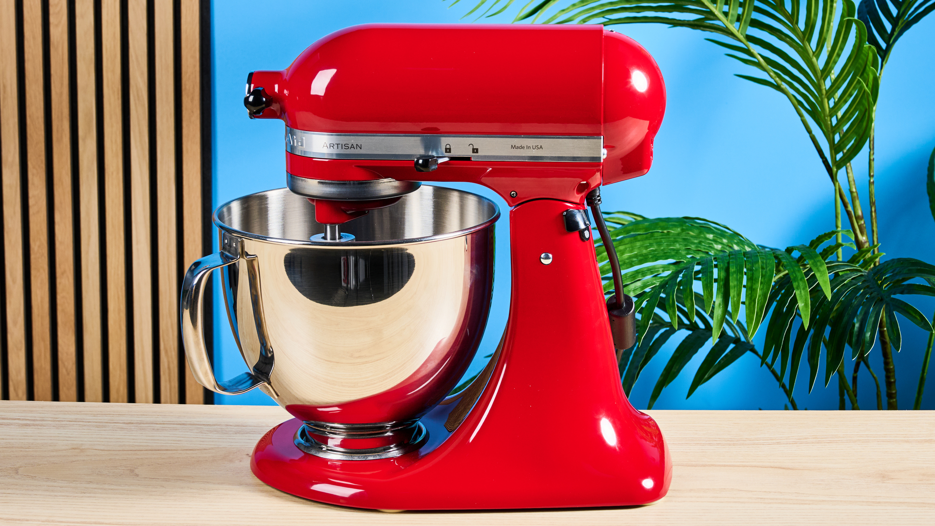 photograph of the kitchenaid artisan stand mixer tilt head in cherry red, with the mixing bowl, paddle, dough hook, and whisk attachments, photographed against a blue background
