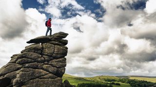 A hiker standing on top of a large rock at Hound Tor in Dartmoor England overlooking a valley