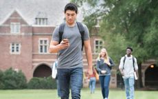 A college student walking across campus checking his phone and wearing a backpack.