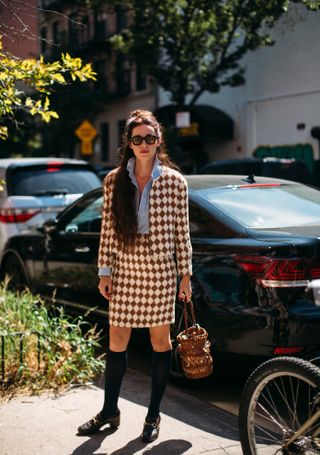 Woman at fashion week wearing preppy outfit featuring an argyle print cardigan, matching skirt, blue shirt, black knee-high socks, and heeled loafers.