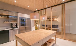 A kitchen with a large wooden table in the middle, a sliver freezer, a fridge with a glass door, an oven, a stove, wall storage shelves and pendant lights.