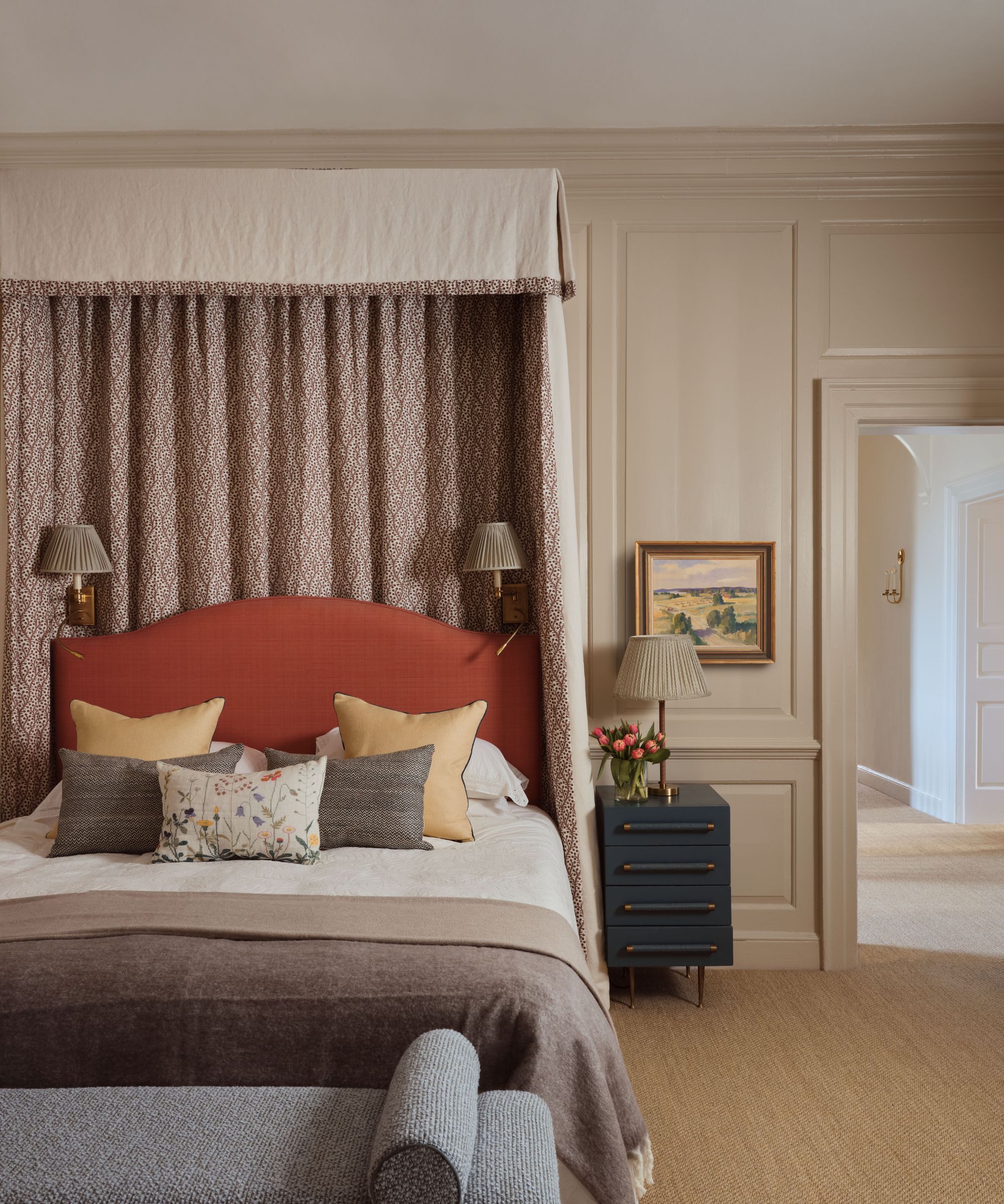 Bedroom with beige painted walls, a canopy bed in a pink fabric, an orange upholstered headboard, yellow and neutral throw pillows, a brown wool throw, and a grey daybed at the end of the bed