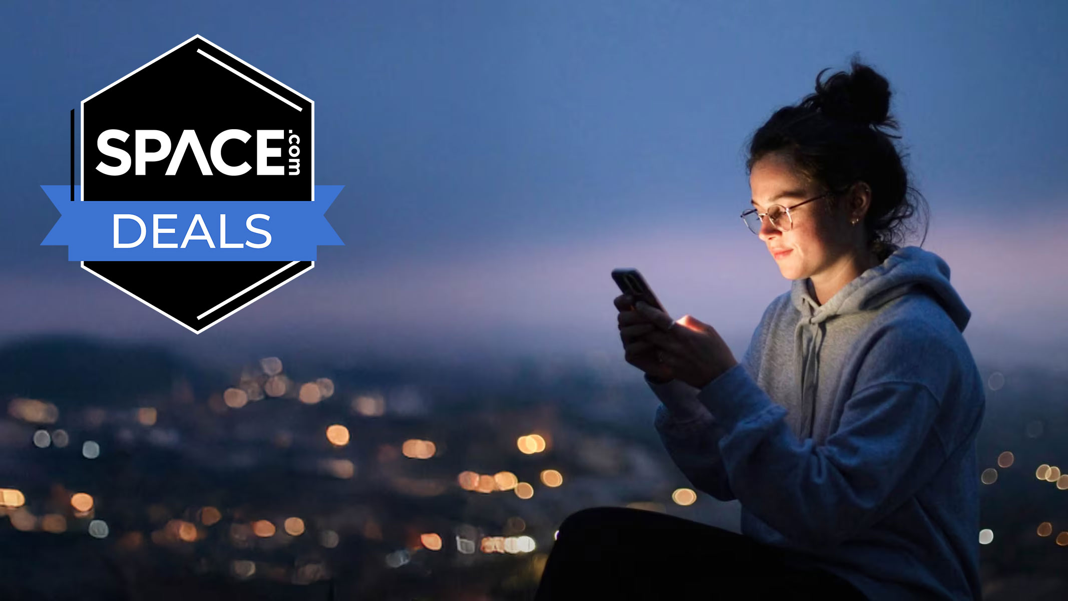 A woman is looking at her mobile phone while wearing a hoodie in front of a city skyline at dusk. The Space.com logo is in the top left corner.
