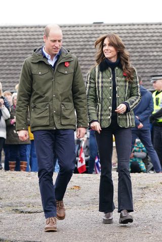 Prince William and Kate Middleton wearing green jackets and brown boots walking on a gravel path outside