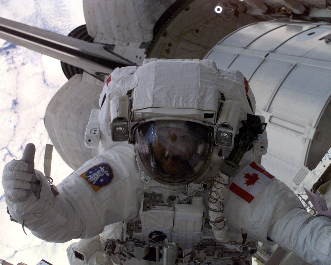 Astronaut Chris Hadfield is pictured in full space gear and suit, doing a thumbs up, whilst floating outside the space station, with the shuttle visible in the background as they hover in space above the cloud covered earth