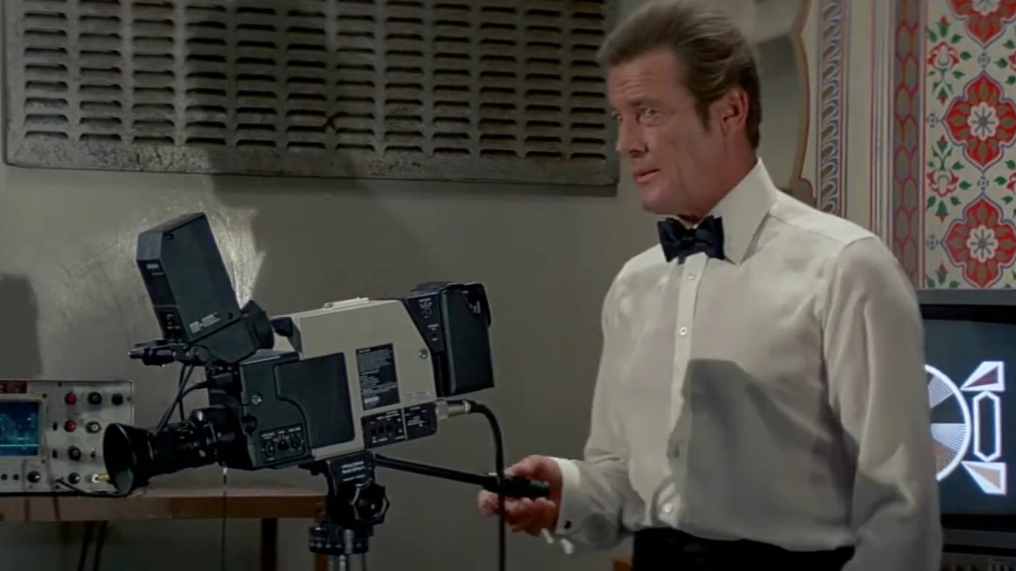 Roger Moore in a white shirt and black bowtie, looking into a television camera.