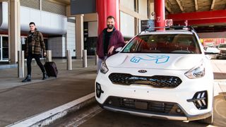 A photo of a tele-operated Kia taxi run by Mapless AI at Pittsburgh International Airport. To the left of the car, a man leans on the car while smiling and a passenger walks past with a suitcase.