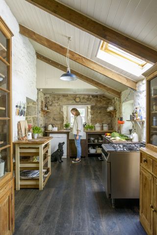 a woman and a dog standing in a free standing kitchen with exposed brick wall, a sloped ceiling with wood beams, and a dark wood floor